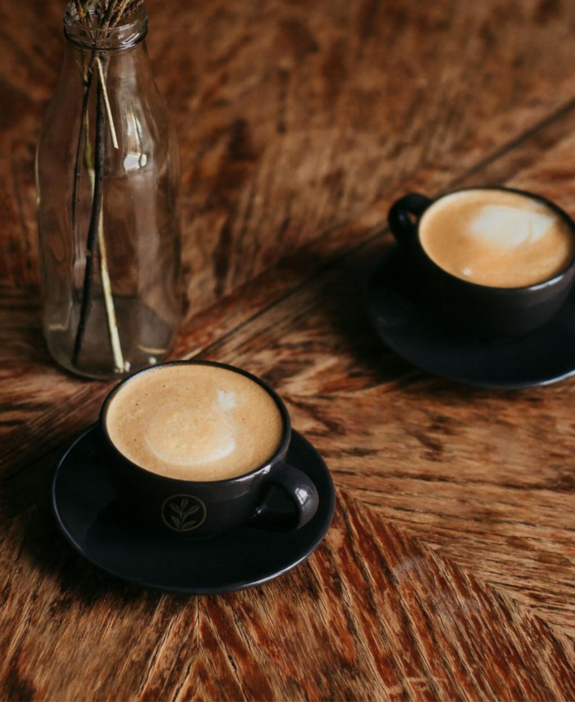 Schwarze Cafe Tassen mit Logo auf braunem Tisch und Deko daneben