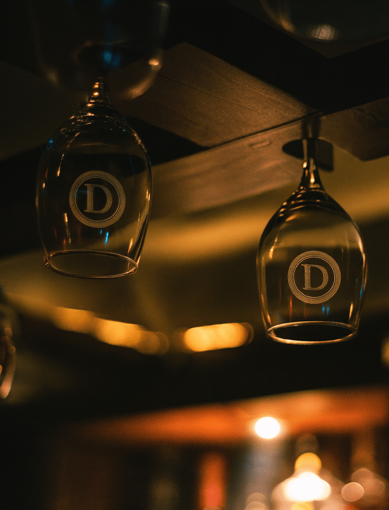 Fokus auf Weingläser, hängen in einer Vorrichtung mit eingravierten Logos und dimmenden Lichtern verschwommen im Hintergrund. Restaurant-Ambiente