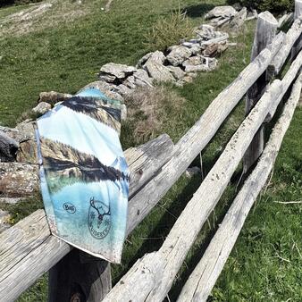 Ein multifunktionales Tuch mit Berg- und See-Motiv hängt über einem alten Holzzaun in einer grünen Wiesenlandschaft. Im Hintergrund sind Steine und Gräser zu sehen