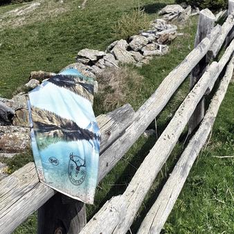 Ein multifunktionales Tuch mit Berg- und See-Motiv hängt über einem alten Holzzaun in einer grünen Wiesenlandschaft. Im Hintergrund sind Steine und Gräser zu sehen