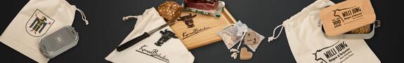 Mehrere nachhaltige Geschenksets liegen nebeneinander: Baumwollbeutel mit Firmenlogo, ein Brotzeitbrett mit Messer und Brot, eine Edelstahl-Brotdose mit Holzdeckel und Aufdruck sowie eine kleine Schafmilchseife im Organzabeutel
