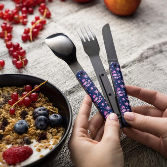 Eine Person hält ein Besteckset aus Messer, Gabel und Löffel mit dunkelblauen Griffen und rosa Blütenmuster in den Händen. Daneben steht eine Schale mit Joghurt, Müsli, Beeren und Obst auf einem Tisch.