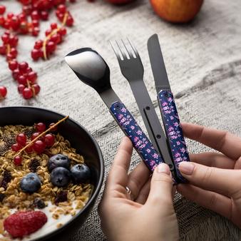 Eine Person hält ein Besteckset aus Messer, Gabel und Löffel mit dunkelblauen Griffen und rosa Blütenmuster in den Händen. Daneben steht eine Schale mit Joghurt, Müsli, Beeren und Obst auf einem Tisch.