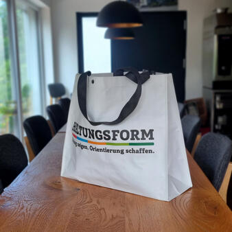Eine weiße Tragetasche mit schwarzen Henkeln steht auf einem Holztisch in einem modernen Besprechungsraum. Auf der Tasche steht ein mehrfarbiger Aufdruck mit dem Wort „HALTUNGSFORM“ und einem Slogan darunter.