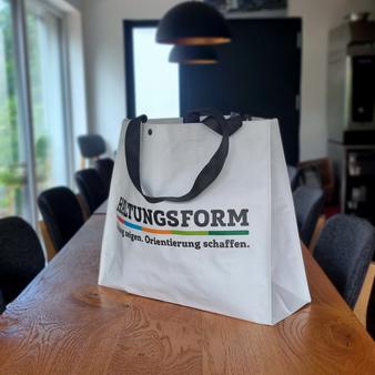 Eine weiße Tragetasche mit schwarzen Henkeln steht auf einem Holztisch in einem modernen Besprechungsraum. Auf der Tasche steht ein mehrfarbiger Aufdruck mit dem Wort „HALTUNGSFORM“ und einem Slogan darunter.