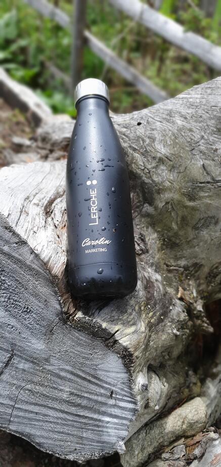 Eine schwarze Trinkflasche aus Edelstahl mit Wassertropfen liegt auf einem verwitterten Baumstamm. Auf der Flasche sind ein vertikales Logo mit der Aufschrift „LERCHE“ sowie der Name „Carolin" und die Abteilung "Marketing“ in Weiß zu sehen