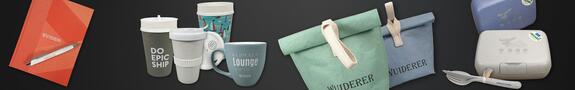Aufgereiht vor dunklem Hintergrund liegen verschiedene nachhaltige Werbeartikel: ein rotes Notizbuch mit Kugelschreiber, mehrere wiederverwendbare Becher und eine Tasse, zwei faltbare Lunchbags in Grün und Blau mit Stoffschlaufen sowie zwei Brotdosen mit passendem Besteck