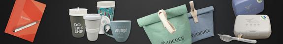 Aufgereiht vor dunklem Hintergrund liegen verschiedene nachhaltige Werbeartikel: ein rotes Notizbuch mit Kugelschreiber, mehrere wiederverwendbare Becher und eine Tasse, zwei faltbare Lunchbags in Grün und Blau mit Stoffschlaufen sowie zwei Brotdosen mit passendem Besteck