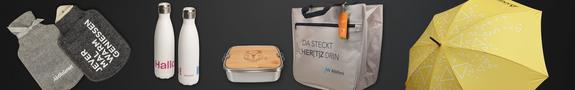 Auf dunklem Hintergrund sind verschiedene Werbeartikel angeordnet: zwei graue Wärmflaschen mit Stoffbezug, zwei weiße Edelstahltrinkflaschen, eine Lunchbox aus Metall mit Bambusdeckel, eine beige Tragetasche mit schwarzem Griff und ein gelber Regenschirm mit geometrischem Muster.