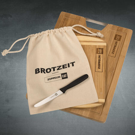 Bambus-Schneidebrett mit Logo liegt zusammen mit einem Stoffbeutel und einem Messer auf grauem Hintergrund