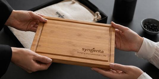 Zwei Personen überreichen sich ein graviertes Holzschneidebrett auf einem Schreibtisch im Büro, im Hintergrund liegt ein Stoffbeutel mit Logo