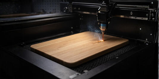 Lasergravur eines Logos auf einem hellen Holz-Schneidebrett in einer modernen Maschine, mit leicht sichtbarem Rauch am Gravurpunkt