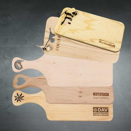 Mehrere Holzschneidebretter mit unterschiedlichen Lasercut-Ausschnitten und gravierten Logos auf grauem Hintergrund