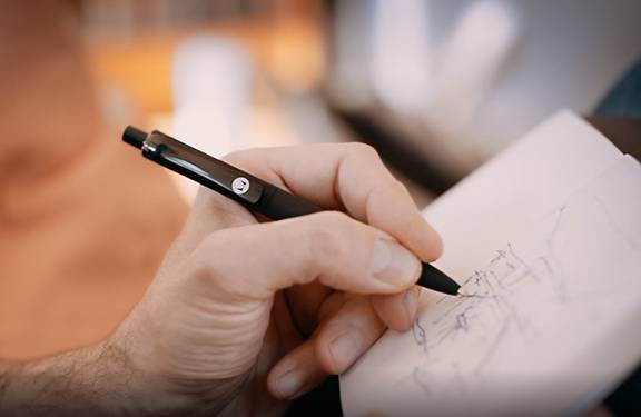 Eine Hand hält einen schwarzen Kugelschreiber und schreibt oder skizziert auf einem weißen Notizblock. Der Fokus liegt auf dem Stift und den Fingern, im Hintergrund ist alles unscharf.