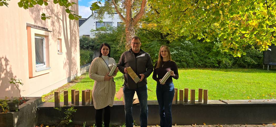 Drei Personen stehen im Freien vor einem Gebäude und halten silberne Thermoflaschen in den Händen. Auf der Mauer vor ihnen sind mehrere gleichartige Kartons aufgereiht. Im Hintergrund ist eine grüne Rasenfläche mit Bäumen und herbstlichem Laub zu sehen.