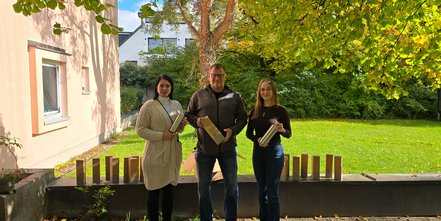 Drei Personen stehen im Freien vor einem Gebäude und halten silberne Thermoflaschen in den Händen. Auf der Mauer vor ihnen sind mehrere gleichartige Kartons aufgereiht. Im Hintergrund ist eine grüne Rasenfläche mit Bäumen und herbstlichem Laub zu sehen.