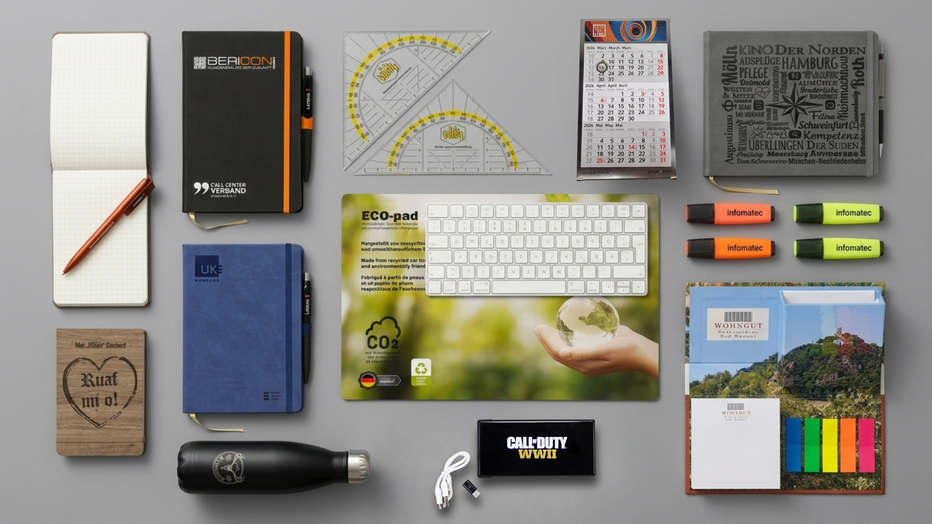 Büroartikel mit Logo liegen auf grauem Hintergrund sortiert nebeneinander und übereinander.