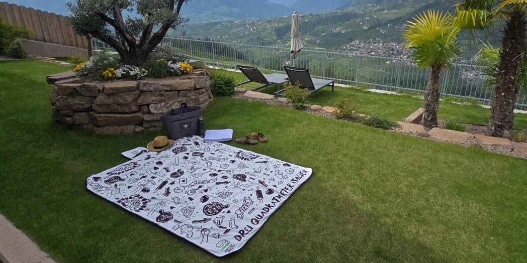Es wird eine große vollflächig bedruckte Picknickdecke mit gustavo Gusto Logo auf einer Hotelanlage auf Wiese gezeigt.