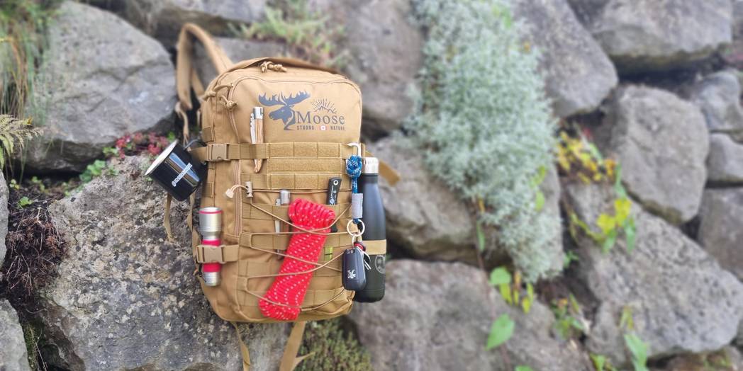 Ein olivbrauner Outdoor-Rucksack mit mehreren Taschen und Befestigungsschlaufen steht auf einer hölzernen Parkbank. Außen sind verschiedene Gegenstände befestigt, darunter eine rote Kordel, eine Taschenlampe, ein schwarzer Metallbecher und Karabiner. Im Hintergrund sind ein Kiesweg und grüne Vegetation zu sehen.