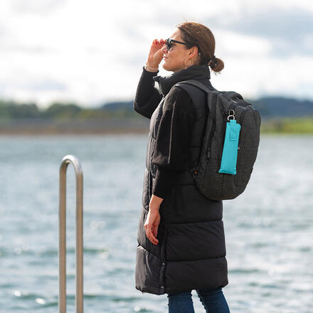 Person steht am Ufer eines Gewässers und blickt in die Ferne. Sie trägt eine lange, schwarze Steppweste, Jeans und eine Sonnenbrille. An ihrem grauen Rucksack hängt ein zusammengefalteter Taschenschirm in einer türkisfarbenen Hülle mit Karabiner.