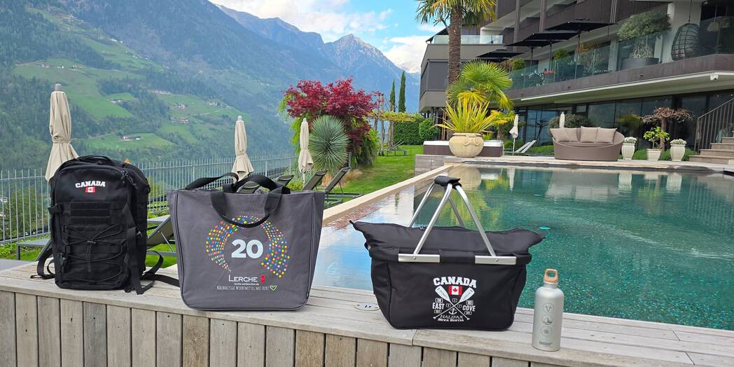 3 verschiedene taschenmodelle stehen an einerm Pool nebeneinander gestellt. Alle sind individuell bedruckt mit Logo und im Hintergrund sieht man eine Hotelanlage und die Berge Südtirols.