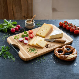 Bambus Schneidebrett mit schwarzem Metallgriff, belegt mit Käse, Radieschen, Tomaten und Brezel auf dunkler Oberfläche