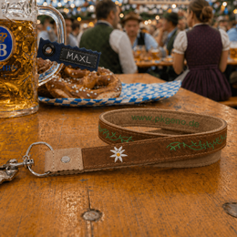 Brauner Leder-Werbeschlüsselanhänger im Trachtenstil mit Edelweiß-Stickerei und grünem Firmenlogo „pkgeno.de“ als Veredelung.