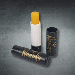 Schwarzer Lippenpflegestift als Werbeartikel mit goldfarbenem Wanuka-Logodruck und gelbem Pflegestick in geöffneter Ausführung.