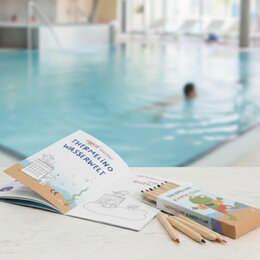 Aufgeschlagenes Malbuch mit Buntstiften auf einem Tisch vor einem Schwimmbecken in einer Therme