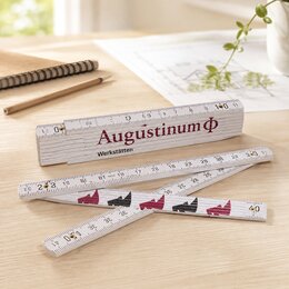 Weißer Holz-Gliedermaßstab mit Augustinum-Logo, teilweise aufgeklappt auf einem hellen Holztisch, daneben Bleistift, Notizbuch und Skizze.