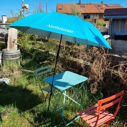 Hellblauer Sonnenschirm mit silber Adelholzener Schriftzug über einem Gartentisch im Außenbereich