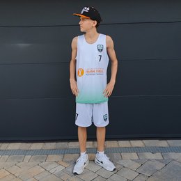 Eine stehende Person ist von vorne zu sehen. Sie trägt ein ärmelloses, helles Sporttrikot mit grünem Farbverlauf am unteren Rand, der Zahl 7 und mehreren Vereins- und Sponsorenlogos auf der Brust. Dazu passen helle Shorts mit einem Vereinslogo und der Zahl 7. Die Person trägt außerdem eine schwarze Kappe mit orangenen Akzenten sowie weiße Sportschuhe und weiße Socken. Sie blickt seitlich nach rechts und steht auf einem gepflasterten Untergrund vor einer dunkelgrauen Wand.