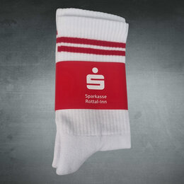 Weiße Sportsocke mit zwei roten Streifen am Sockenbund in Papierbanderole mit Sparkassen-Logo gefaltet auf grauem Hintergrund fotografiert