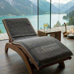 Graues Waffel-Duschtuch als Werbeartikel aus Baumwolle mit eingeprägtem Firmenlogo „Seehotel Einwaller Achensee“ auf Holzliege im Boutiquehotel mit Bergblick.
