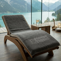 Graues Waffel-Duschtuch als Werbeartikel aus Baumwolle mit eingeprägtem Firmenlogo „Seehotel Einwaller Achensee“ auf Holzliege im Boutiquehotel mit Bergblick.