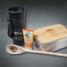 Merchartikel als Nahaufnahme: Edelstahl-Lunchbox und Bambusdeckel mit Lasergravur, Holzlöffel und schwarzem Thermobecher mit Logo Kloster Bräustüberl Reutberg.