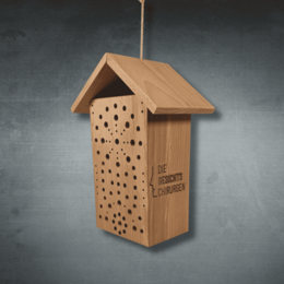 Holz-Wildbienenhotel als Werbeartikel mit Lasergravur und Firmenlogo „Die Gesichts Chirurgen“, aufgehängt vor grauem Hintergrund.