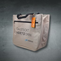 Beigefarbene Werbe-Einkaufstasche aus Stoff, mit schwarzem Reißverschlussfach vorne, schwarzen Henkeln und Aufdruck „Da steckt Her[t]z drin“ 