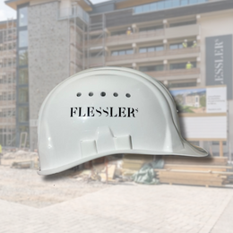 Bauhelm seitlich gezeigt mit einem Domingdruck für das Hotel Flessler. Im Hintergrund eine Baustelle des Hotels zu sehen.