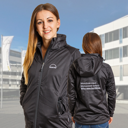 Aufgesticktes Logo auf der Brust und auf dem Rücken einer Softshelljacke zu sehen. Im Hintergrund das Gebäude der MAN: