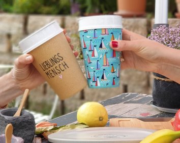 Ein hoher, weißer Trinkbecher mit Henkel ist zu sehen. Auf der Vorderseite befindet sich ein rotes Logo mit Schriftzug. Der Becher hat einen weißen Deckel mit integrierter Trinköffnung und herausnehmbarem Trinkhalm. Der untere Teil ist leicht ergonomisch geformt.