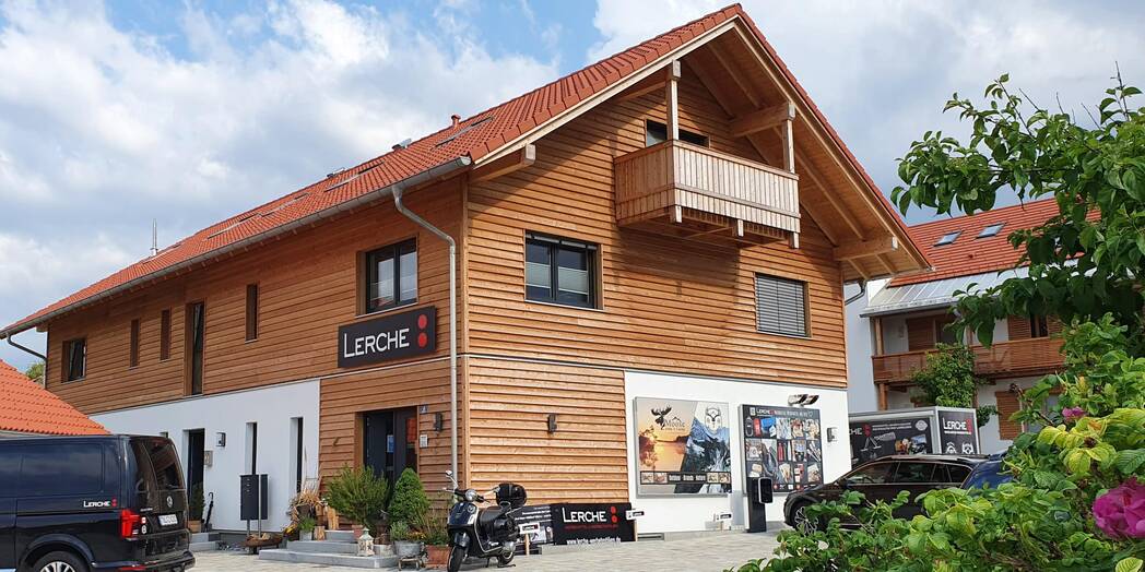 Zweigeschossiges Firmengebäude mit Holzfassade, rotem Ziegeldach, Eingangstür, parkendem Firmenfahrzeug und Lerche Logo an der Außenwand.