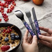 Zwei Hände halten ein dreiteiliges Besteckset aus Messer, Gabel und Löffel mit blauen Griffen, die rosa Blütenmuster zeigen. Im Hintergrund steht eine Schale mit Joghurt, Müsli und Beeren.